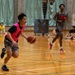 A basketball game or practice session is taking place on an indoor hardwood court. A player wearing a pink vest numbered '9' is dribbling the ball while a taller player in a gray uniform follows behind. Several spectators are seated along the sidelines in what appears to be a gymnasium.