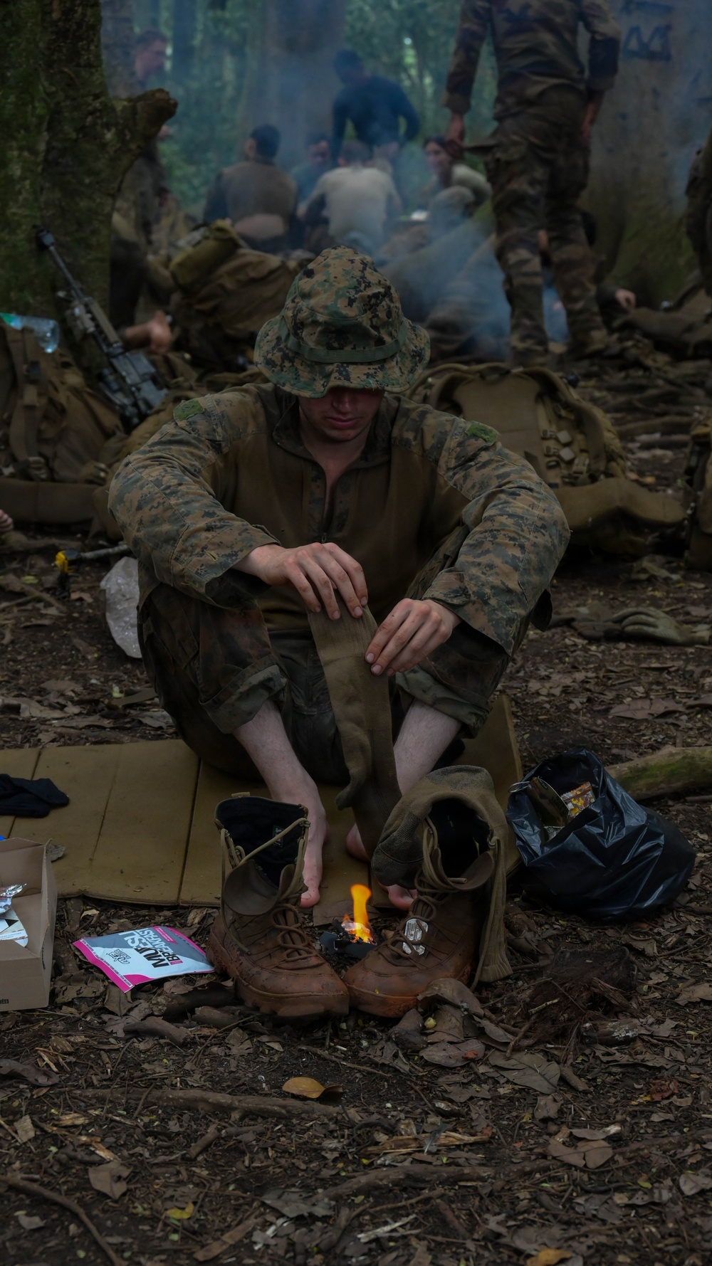 A soldier in MARPAT woodland camouflage uniform and boonie hat sits on the forest floor during what appears to be a field training exercise or field operation. He has removed his boots and socks and is using a small portable stove or heat tab to warm something, likely heating a meal or drying gear. In the background, multiple other soldiers are visible among trees with smoke rising, suggesting a temporary bivouac or rest halt.