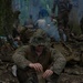 A soldier in MARPAT woodland camouflage uniform and boonie hat sits on the forest floor during what appears to be a field training exercise or field operation. He has removed his boots and socks and is using a small portable stove or heat tab to warm something, likely heating a meal or drying gear. In the background, multiple other soldiers are visible among trees with smoke rising, suggesting a temporary bivouac or rest halt.