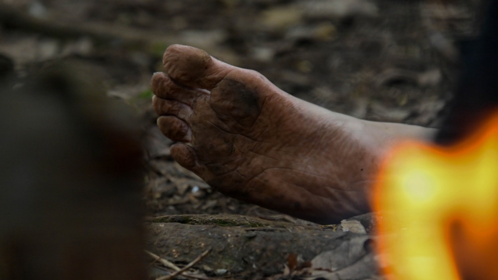 A close-up photograph of a bare, dirty human foot resting on rocky ground in an outdoor setting. The foot appears heavily soiled with dirt and grime, suggesting prolonged barefoot activity outdoors. A blurred flame or fire source is visible in the foreground right side of the frame.
