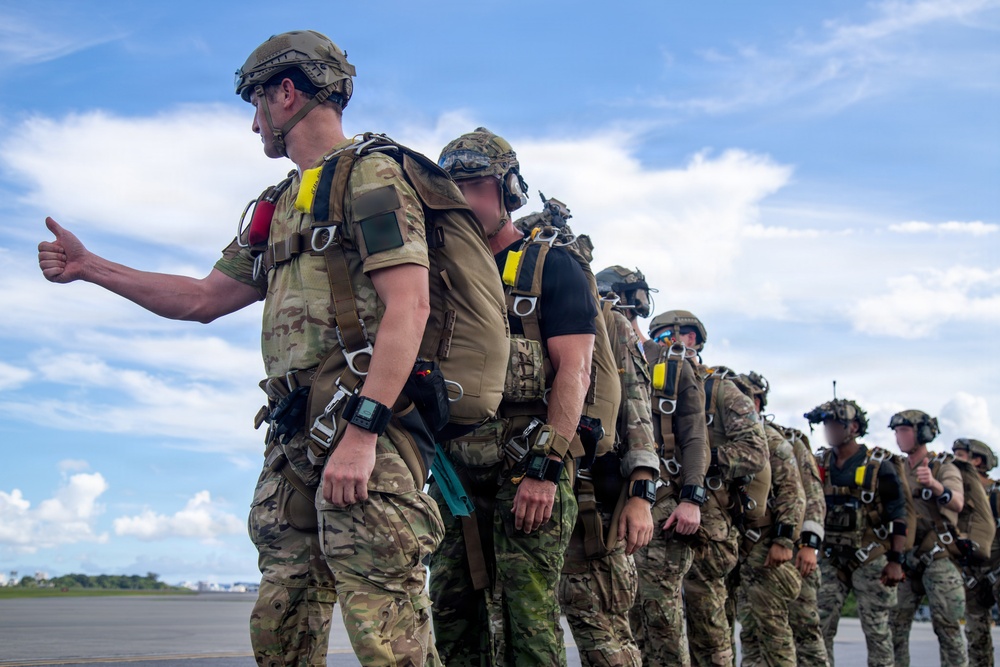 A line of military personnel in full tactical gear, including parachute rigs and combat helmets, are staged on what appears to be an airfield tarmac. The lead soldier is giving a thumbs-up gesture, suggesting readiness or a pre-jump check. The group appears to be preparing for a military parachute operation, likely HALO or HAHO based on the visible oxygen equipment.