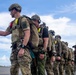 A line of military personnel in full tactical gear, including parachute rigs and combat helmets, are staged on what appears to be an airfield tarmac. The lead soldier is giving a thumbs-up gesture, suggesting readiness or a pre-jump check. The group appears to be preparing for a military parachute operation, likely HALO or HAHO based on the visible oxygen equipment.