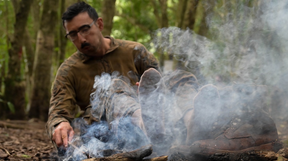 A man wearing glasses and olive/camouflage clothing sits on a forest floor surrounded by smoke, with his feet extended toward what appears to be a smoldering fire or smoke source. His boots are removed and placed nearby, and he appears to be warming or drying his socked feet. The setting is a dense woodland environment with leaf litter on the ground.