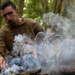 A man wearing glasses and olive/camouflage clothing sits on a forest floor surrounded by smoke, with his feet extended toward what appears to be a smoldering fire or smoke source. His boots are removed and placed nearby, and he appears to be warming or drying his socked feet. The setting is a dense woodland environment with leaf litter on the ground.