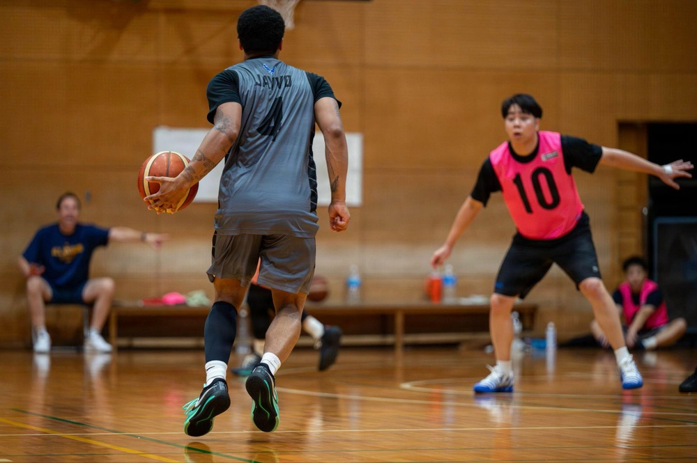 A basketball game is being played in an indoor gymnasium with a wooden court floor. A player wearing a gray jersey with the name 'JAYVO' and number 4 is handling the ball while being defended by a player in a pink vest numbered 10. Additional players and a spectator are visible in the background.