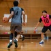 A basketball game is being played in an indoor gymnasium with a wooden court floor. A player wearing a gray jersey with the name 'JAYVO' and number 4 is handling the ball while being defended by a player in a pink vest numbered 10. Additional players and a spectator are visible in the background.