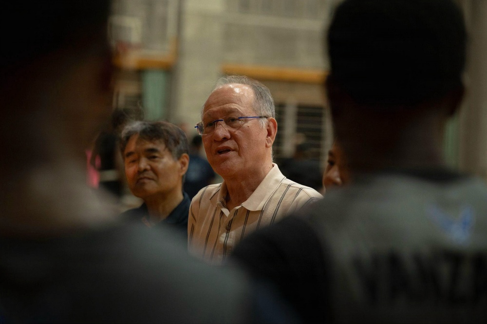 The image shows an older Asian man wearing glasses and a striped collared shirt, appearing to speak or address a group of people. He is surrounded by other individuals, with two figures in the foreground shown from behind in soft focus. The setting appears to be an indoor gymnasium or sports facility.