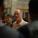The image shows an older Asian man wearing glasses and a striped collared shirt, appearing to speak or address a group of people. He is surrounded by other individuals, with two figures in the foreground shown from behind in soft focus. The setting appears to be an indoor gymnasium or sports facility.