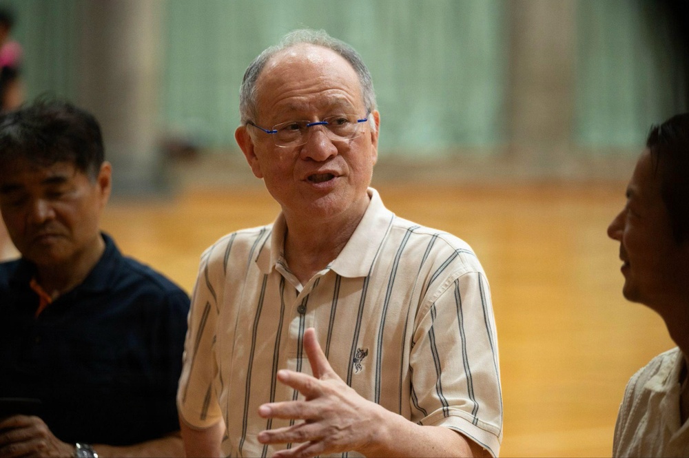 An elderly man wearing blue-framed glasses and a striped polo shirt appears to be speaking or explaining something, with his hand raised in a gesturing motion. He is surrounded by at least two other individuals in what appears to be an indoor sports facility or gymnasium. The image is well-lit and captures what seems to be a casual conversation or discussion.