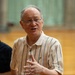 An elderly man wearing blue-framed glasses and a striped polo shirt appears to be speaking or explaining something, with his hand raised in a gesturing motion. He is surrounded by at least two other individuals in what appears to be an indoor sports facility or gymnasium. The image is well-lit and captures what seems to be a casual conversation or discussion.