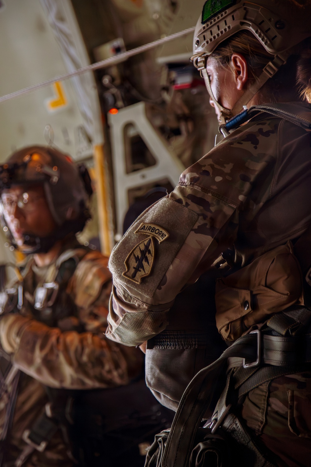 Two military personnel are inside what appears to be a military transport aircraft, likely preparing for an airborne operation. The foreground soldier wears a MultiCam uniform with a clearly visible 'Airborne' tab and Special Forces patch on the left sleeve, along with a tactical helmet and protective eyewear. The setting and equipment suggest a pre-jump or in-flight airborne insertion scenario.