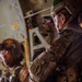 Two military personnel are inside what appears to be a military transport aircraft, likely preparing for an airborne operation. The foreground soldier wears a MultiCam uniform with a clearly visible 'Airborne' tab and Special Forces patch on the left sleeve, along with a tactical helmet and protective eyewear. The setting and equipment suggest a pre-jump or in-flight airborne insertion scenario.