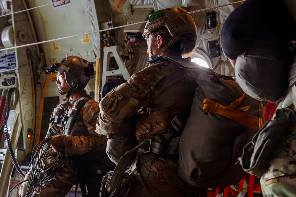 Military personnel in full combat gear and helmets are seated inside a military transport aircraft, appearing to be preparing for a parachute jump operation. The soldiers are equipped with parachute packs, tactical helmets, and multicam uniforms. The interior of the aircraft shows typical military cargo aircraft features including static lines, yellow safety markings, and instrumentation panels.