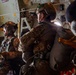 Military personnel in full combat gear and helmets are seated inside a military transport aircraft, appearing to be preparing for a parachute jump operation. The soldiers are equipped with parachute packs, tactical helmets, and multicam uniforms. The interior of the aircraft shows typical military cargo aircraft features including static lines, yellow safety markings, and instrumentation panels.