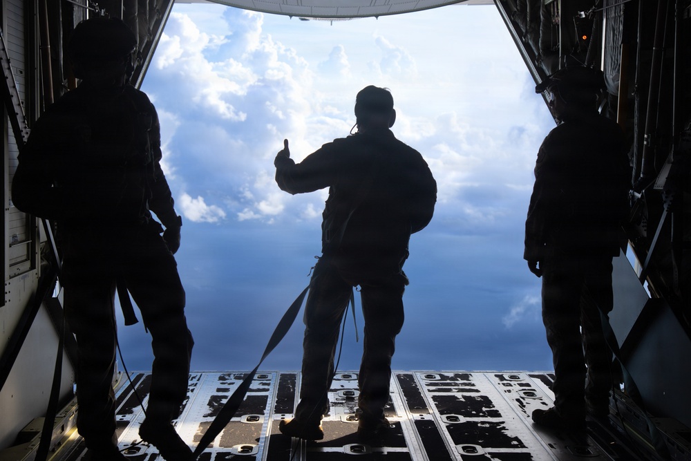 Three silhouetted military personnel stand at the open rear cargo ramp of a military transport aircraft in flight. The central figure appears to be giving a hand signal, possibly a thumbs-up, while the others stand on either side. The scene is backlit by a dramatic sky with cumulus clouds, creating strong silhouette contrast.