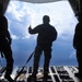 Three silhouetted military personnel stand at the open rear cargo ramp of a military transport aircraft in flight. The central figure appears to be giving a hand signal, possibly a thumbs-up, while the others stand on either side. The scene is backlit by a dramatic sky with cumulus clouds, creating strong silhouette contrast.