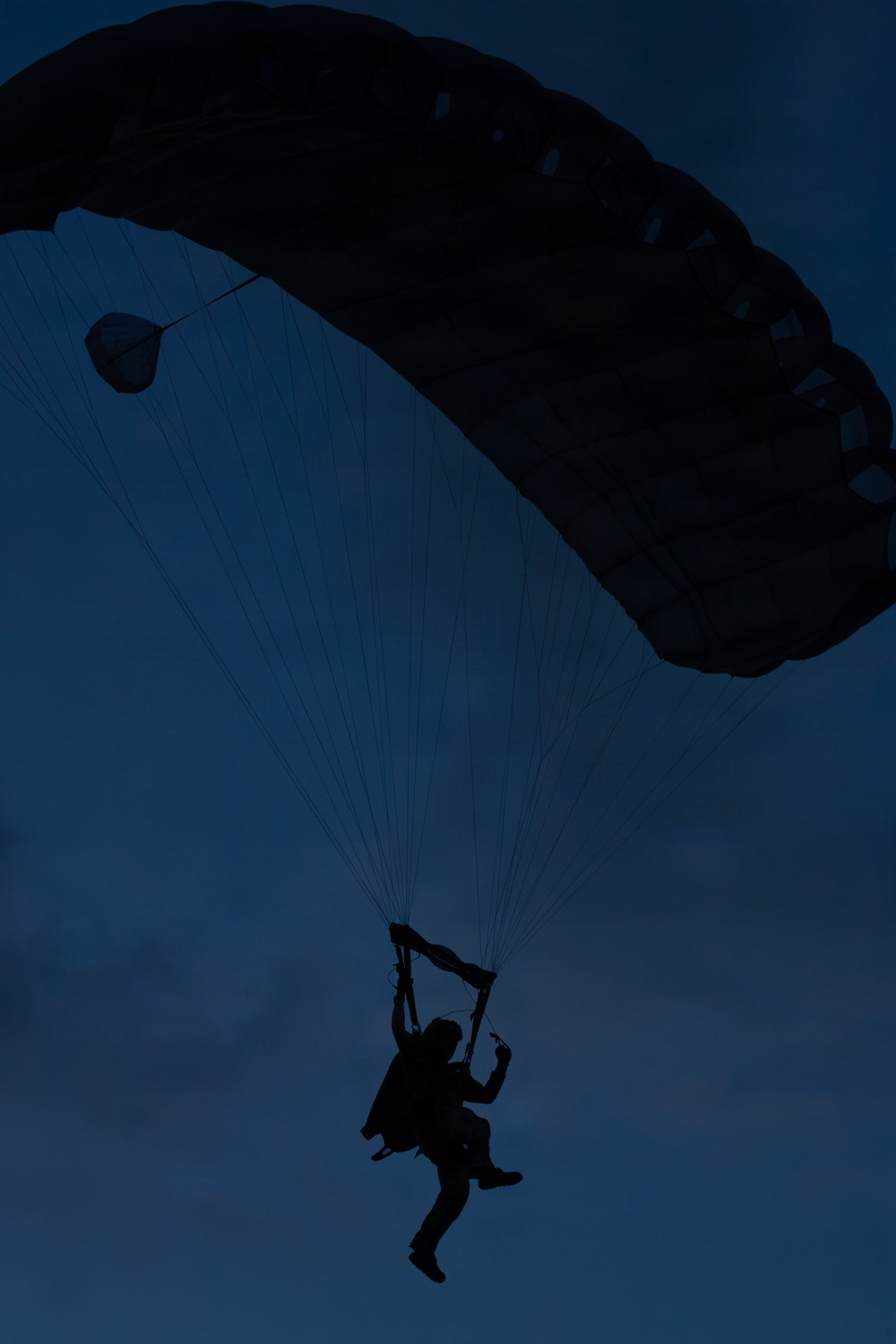 A silhouetted parachutist is captured mid-descent against a deep blue twilight sky. The ram-air parachute canopy is fully deployed and visible in the upper portion of the frame. The low-light conditions render the subject and canopy as dark silhouettes with minimal detail.