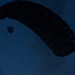 A silhouetted parachutist is captured mid-descent against a deep blue twilight sky. The ram-air parachute canopy is fully deployed and visible in the upper portion of the frame. The low-light conditions render the subject and canopy as dark silhouettes with minimal detail.