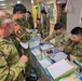 Military personnel from multiple nations are participating in a calligraphy cultural exchange activity, practicing Japanese kanji writing with brushes and ink. The scene appears to take place at a joint military event or exercise, with soldiers wearing Australian, American, and Japanese Self-Defense Force uniforms. A 'Calligraphy' station sign and a 'KANJI 2500' reference book are visible on the table.