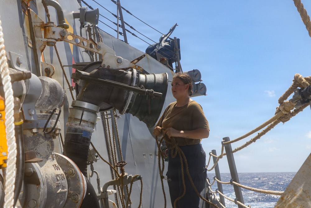 A female sailor or naval personnel stands on the deck of a military or coast guard vessel at sea, handling rope lines. She is positioned alongside large industrial piping and valve equipment mounted on the ship's hull. The ocean is visible in the background under clear blue skies.