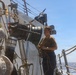 A female sailor or naval personnel stands on the deck of a military or coast guard vessel at sea, handling rope lines. She is positioned alongside large industrial piping and valve equipment mounted on the ship's hull. The ocean is visible in the background under clear blue skies.