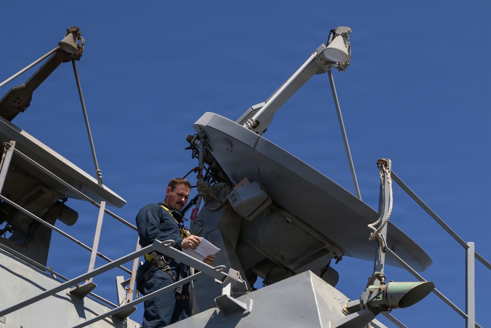 A naval technician or sailor in a blue uniform and safety harness is standing on an elevated platform of a military vessel, reviewing documents near a large satellite or radar dish antenna. The image is taken from a low angle looking upward against a clear blue sky. The setting appears to be aboard a warship's superstructure during a maintenance or inspection activity.