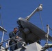 A naval technician or sailor in a blue uniform and safety harness is standing on an elevated platform of a military vessel, reviewing documents near a large satellite or radar dish antenna. The image is taken from a low angle looking upward against a clear blue sky. The setting appears to be aboard a warship's superstructure during a maintenance or inspection activity.