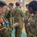 A multinational military gathering showing personnel from at least three nations — Japan, Australia, and the United States — engaged in what appears to be a joint training or coordination exercise indoors. A Japanese soldier wearing an orange 'Interpreter' vest (通訳) is interacting with a U.S. Army soldier bearing an 11th Airborne Division patch. Australian soldiers with flag patches are visible in the background.