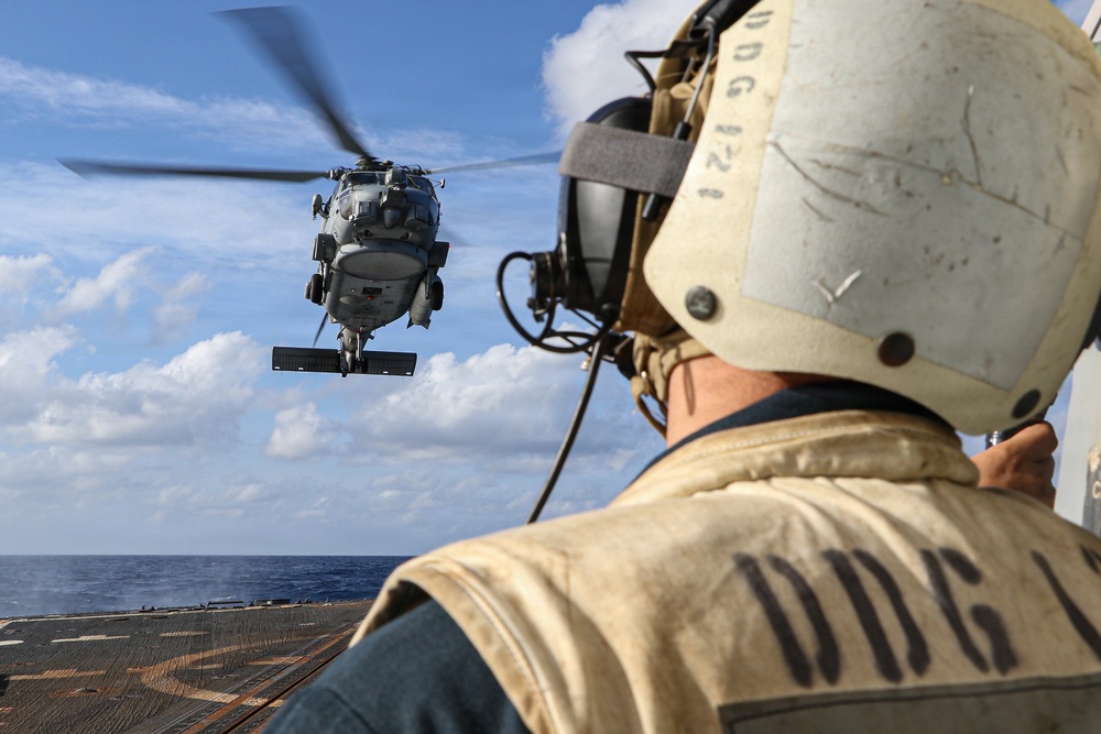A naval flight deck crew member wearing a protective helmet and vest marked 'DDG' observes a military helicopter hovering above the flight deck of a U.S. Navy destroyer at sea. The helicopter appears to be in the process of landing or departing from the ship's flight deck. The scene takes place under partly cloudy skies over open ocean.