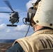 A naval flight deck crew member wearing a protective helmet and vest marked 'DDG' observes a military helicopter hovering above the flight deck of a U.S. Navy destroyer at sea. The helicopter appears to be in the process of landing or departing from the ship's flight deck. The scene takes place under partly cloudy skies over open ocean.