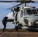 A military helicopter, identified by the number '51' on its fuselage, is secured on the flight deck of a naval vessel with chains and tie-downs. A deck crew member in blue coveralls and hearing protection is leaning forward, likely performing a pre-flight check or securing procedure. The rotors are spinning, indicating the aircraft is either preparing for takeoff or has recently landed.
