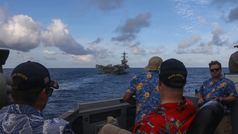 Several individuals wearing 'Black Knights' caps and Hawaiian shirts observe a large aircraft carrier from what appears to be a smaller vessel at sea. The scene suggests a naval gathering or transit operation, with the carrier visible in the middle distance against a partly cloudy sky. The casual attire combined with military-branded caps suggests this may be a Tiger Cruise or similar informal naval event.