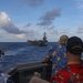 Several individuals wearing 'Black Knights' caps and Hawaiian shirts observe a large aircraft carrier from what appears to be a smaller vessel at sea. The scene suggests a naval gathering or transit operation, with the carrier visible in the middle distance against a partly cloudy sky. The casual attire combined with military-branded caps suggests this may be a Tiger Cruise or similar informal naval event.
