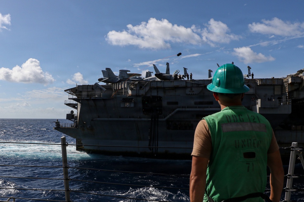 A U.S. Navy sailor wearing a green hard hat and green vest marked 'DDG 121 UNREP 71' observes a large aircraft carrier during an underway replenishment (UNREP) operation at sea. Multiple fixed-wing aircraft, including what appear to be F/A-18 Hornets/Super Hornets and EA-18G Growlers, are visible on the carrier's flight deck. A transfer package or line appears to be mid-air between the two vessels, consistent with a connected replenishment evolution.