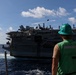 A U.S. Navy sailor wearing a green hard hat and green vest marked 'DDG 121 UNREP 71' observes a large aircraft carrier during an underway replenishment (UNREP) operation at sea. Multiple fixed-wing aircraft, including what appear to be F/A-18 Hornets/Super Hornets and EA-18G Growlers, are visible on the carrier's flight deck. A transfer package or line appears to be mid-air between the two vessels, consistent with a connected replenishment evolution.
