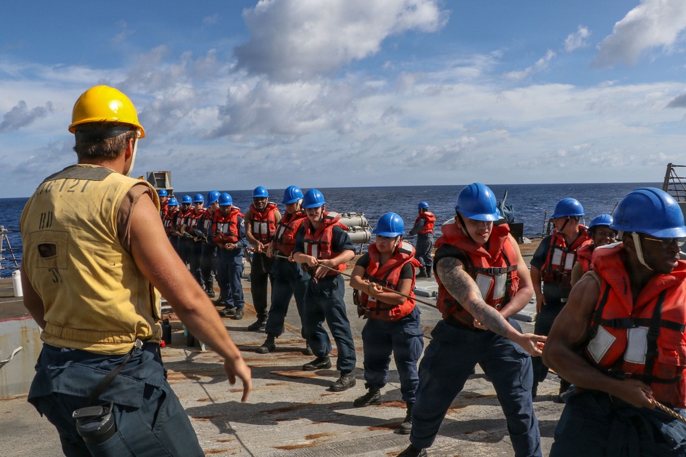 A group of naval personnel are conducting a line-handling or replenishment-at-sea (RAS) drill on the deck of a military vessel at sea. Sailors wearing blue hard hats and orange life vests are pulling on a rope or line under the supervision of a supervisor wearing a yellow hard hat and tan vest. The exercise appears to be a coordinated training evolution taking place in open ocean conditions.