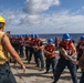 A group of naval personnel are conducting a line-handling or replenishment-at-sea (RAS) drill on the deck of a military vessel at sea. Sailors wearing blue hard hats and orange life vests are pulling on a rope or line under the supervision of a supervisor wearing a yellow hard hat and tan vest. The exercise appears to be a coordinated training evolution taking place in open ocean conditions.