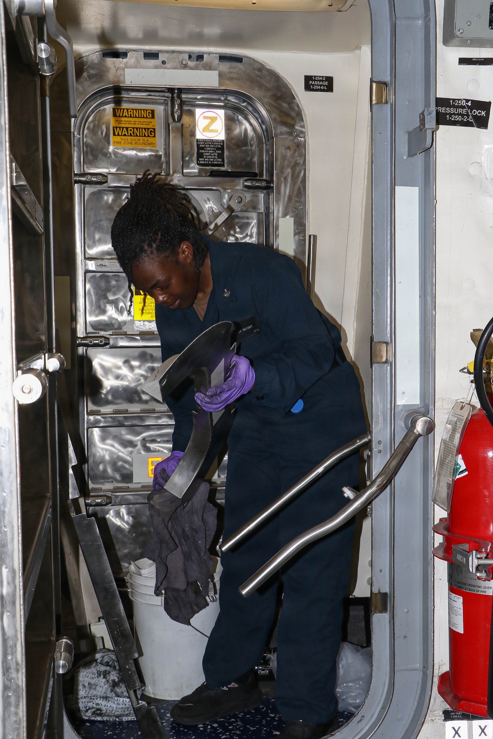 A Navy sailor in dark blue coveralls and purple nitrile gloves is cleaning or polishing metal components near a watertight door aboard a naval vessel. The sailor is working in a passageway area with a white bucket and dark cleaning cloths. The environment shows typical naval ship interior features including warning placards, zone markings, and a fire extinguisher.