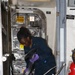 A Navy sailor in dark blue coveralls and purple nitrile gloves is cleaning or polishing metal components near a watertight door aboard a naval vessel. The sailor is working in a passageway area with a white bucket and dark cleaning cloths. The environment shows typical naval ship interior features including warning placards, zone markings, and a fire extinguisher.