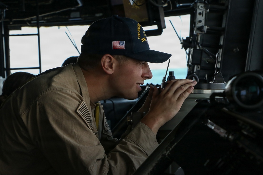 A U.S. Navy sailor is stationed at what appears to be a bridge wing or navigation station aboard a naval vessel. The sailor is wearing a ship's ball cap with 'USS' and 'DDG' visible, indicating a guided-missile destroyer, and is speaking into a handheld radio while operating navigational equipment. The image captures the sailor in an active duty role performing bridge watch or navigation duties.