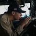 A U.S. Navy sailor is stationed at what appears to be a bridge wing or navigation station aboard a naval vessel. The sailor is wearing a ship's ball cap with 'USS' and 'DDG' visible, indicating a guided-missile destroyer, and is speaking into a handheld radio while operating navigational equipment. The image captures the sailor in an active duty role performing bridge watch or navigation duties.