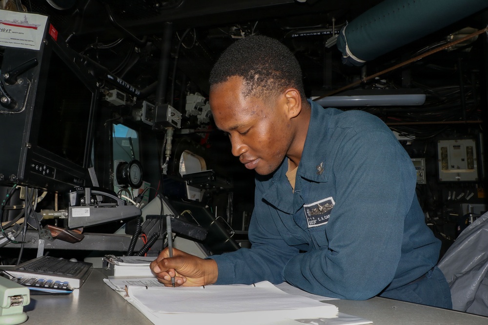 A U.S. Navy sailor in blue coveralls is writing on documents at a workstation aboard what appears to be a naval vessel. The individual is leaning over a desk covered with communications equipment, logbooks, and electronic devices. The setting appears to be a ship's bridge or combat information center.