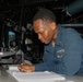 A U.S. Navy sailor in blue coveralls is writing on documents at a workstation aboard what appears to be a naval vessel. The individual is leaning over a desk covered with communications equipment, logbooks, and electronic devices. The setting appears to be a ship's bridge or combat information center.