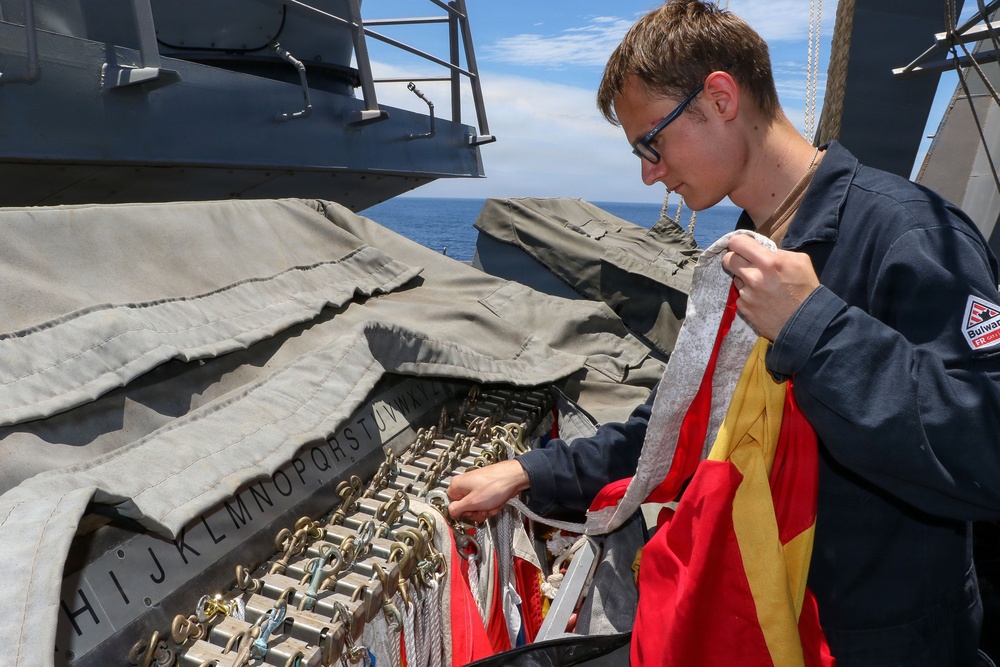A young naval personnel member wearing a dark blue uniform with a 'Bulwark' patch is handling signal flags on the deck of a warship at sea. They are working with a flag locker or signal flag storage rack labeled with alphabetical letters (H through W visible), which holds multiple clips and halyards for flag hoisting. The ocean is visible in the background under a clear blue sky.