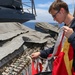 A young naval personnel member wearing a dark blue uniform with a 'Bulwark' patch is handling signal flags on the deck of a warship at sea. They are working with a flag locker or signal flag storage rack labeled with alphabetical letters (H through W visible), which holds multiple clips and halyards for flag hoisting. The ocean is visible in the background under a clear blue sky.