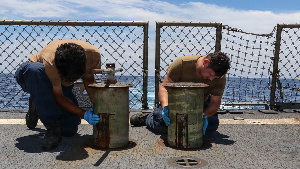 Two military personnel in tan/olive uniforms are performing maintenance work on what appear to be deck fittings or bollards aboard a naval vessel at sea. Both individuals are wearing blue protective gloves and are kneeling or crouching on the ship's deck. The ocean is visible in the background beyond a chain-link safety fence.