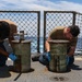 Two military personnel in tan/olive uniforms are performing maintenance work on what appear to be deck fittings or bollards aboard a naval vessel at sea. Both individuals are wearing blue protective gloves and are kneeling or crouching on the ship's deck. The ocean is visible in the background beyond a chain-link safety fence.