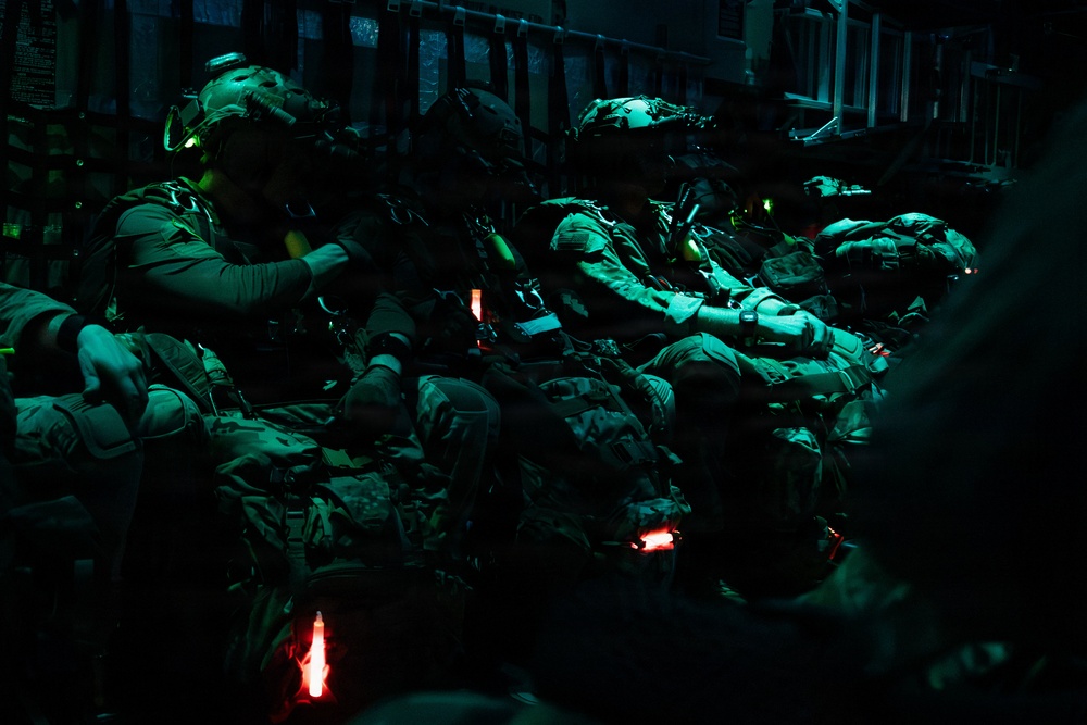 A group of heavily equipped military personnel are seated inside what appears to be a military transport aircraft, illuminated by green and red chemical light sticks (chemlights). The soldiers are wearing full tactical gear including helmets with night vision device mounts, body armor, and carrying large rucksacks. The dark, low-light environment and equipment configuration suggest a pre-jump or insertion preparation scenario.
