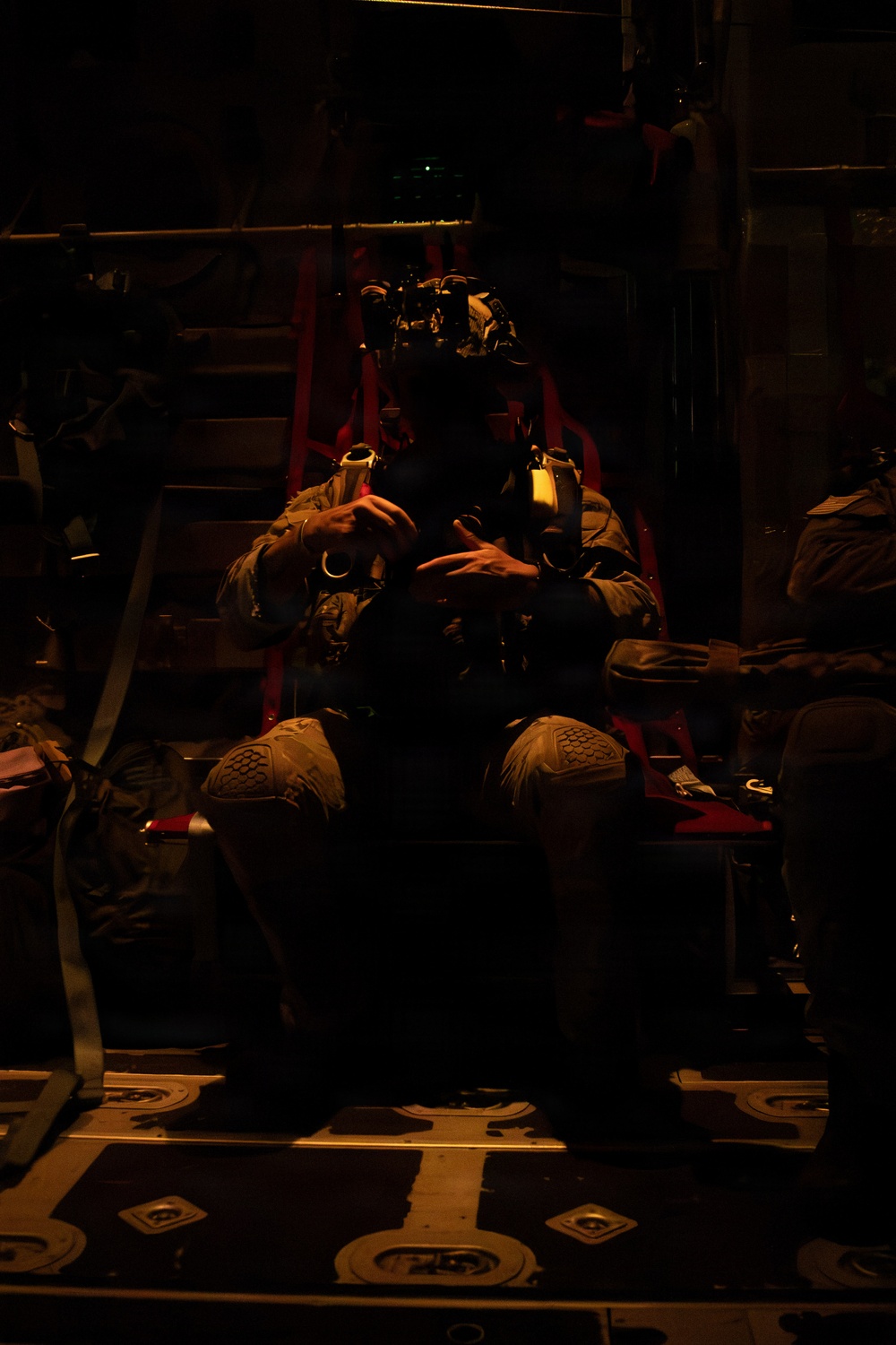 A military operator is seated inside what appears to be a large military transport aircraft, illuminated by dim red/amber lighting. The individual is wearing full tactical gear including a helmet mounted with night vision goggles (NVGs) and a chest harness/rig. The scene suggests a pre-mission or in-flight posture during a nighttime operation.
