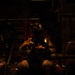 A military operator is seated inside what appears to be a large military transport aircraft, illuminated by dim red/amber lighting. The individual is wearing full tactical gear including a helmet mounted with night vision goggles (NVGs) and a chest harness/rig. The scene suggests a pre-mission or in-flight posture during a nighttime operation.